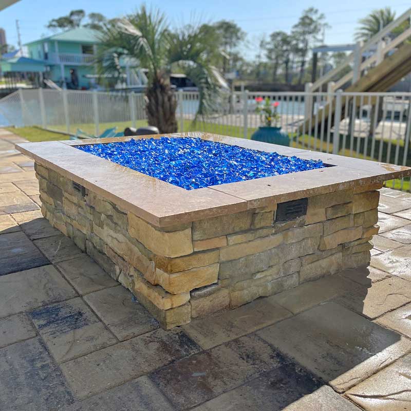 custom gas stone firepit on stone patio filled with cobalt blue glass with palm trees in background